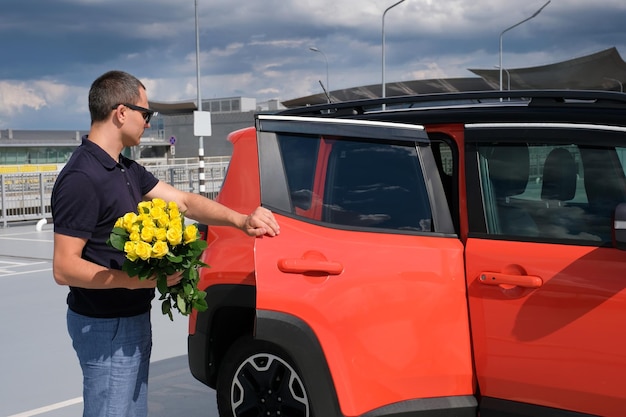 A man with a bouquet of yellow roses opens the door of his car in the parking lot Travel concept