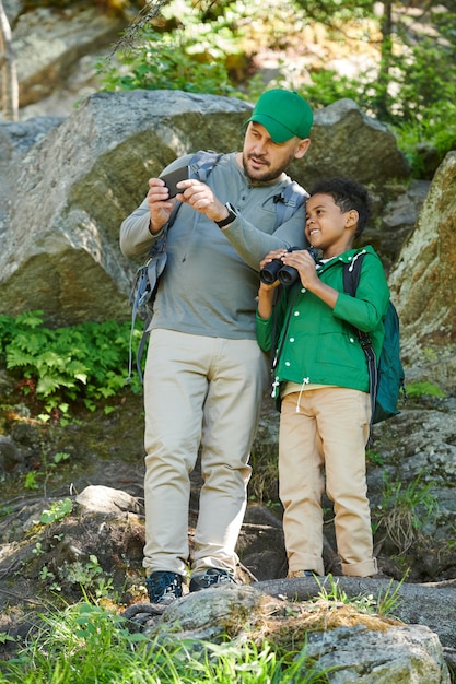 Photo man with the boy standing on the rocks and taking picture of beautiful nature on mobile phone