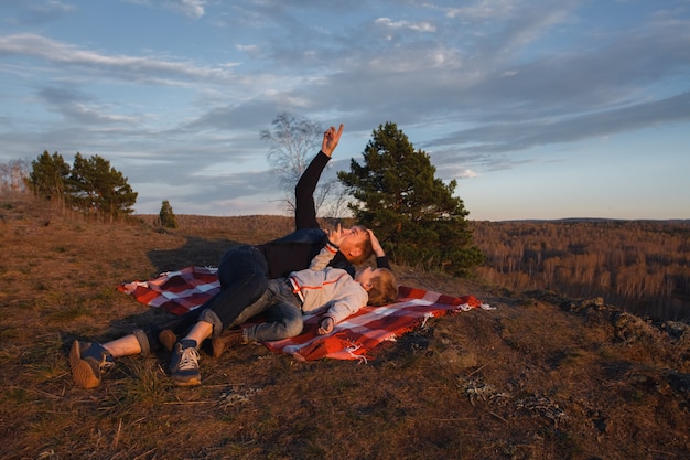 A man with a child lying on a red blanket in the mountains and look at the sky.