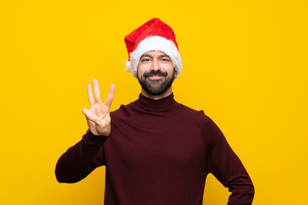 Man with christmas hat over isolated yellow wall happy and counting three with fingers