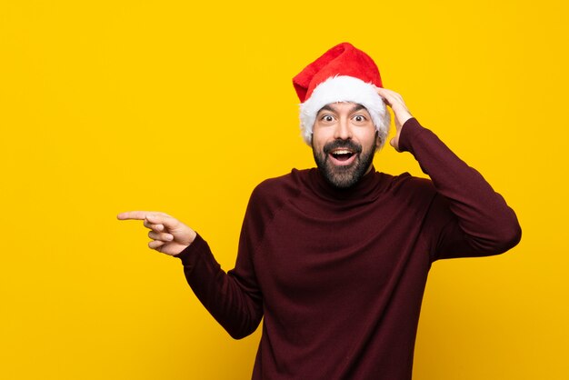 Photo man with christmas hat over isolated yellow wall surprised and pointing finger to the side