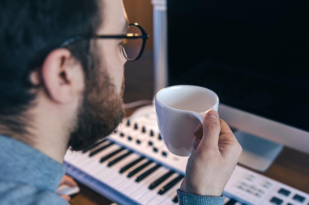 A man with a cup of coffee sits in front of a computer and a musical keyboard