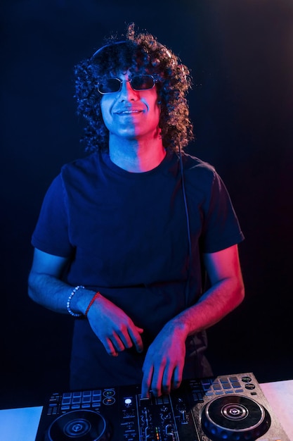 Man with curly hair using DJ equipment and standing in the dark neon lighted room