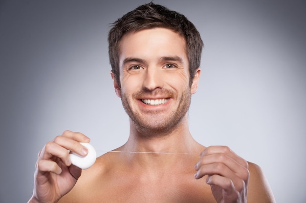 Man with dental floss. Portrait of handsome young shirtless man holding dental floss and smiling while isolated on grey background