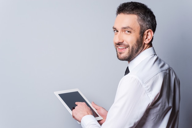 Uomo con tavoletta digitale. vista posteriore di un uomo maturo felice in camicia e cravatta che lavora su un tablet digitale e si guarda alle spalle mentre si trova in piedi su uno sfondo grigio