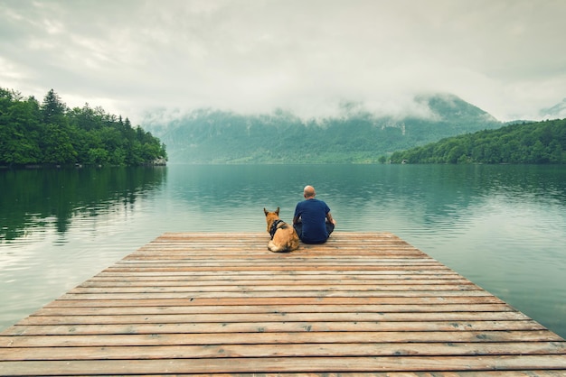 Photo man with dog sitting at wooden pier at bohinj lake slovenia