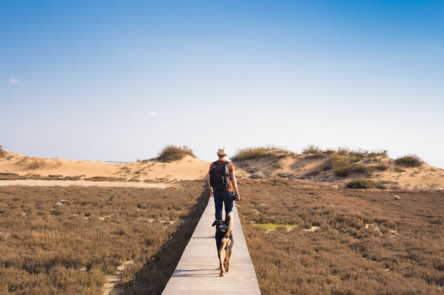 Uomo con cane che cammina sul sentiero di legno sulla spiaggia e guarda in lontananza l'oceano.