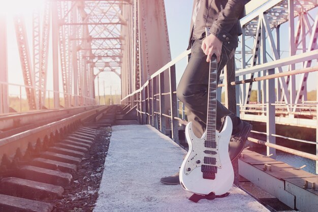 A man with an electric guitar on the railway. A musician in a leather jacket with a guitar on the street in the industrial zone. Guitarist on the bridge.