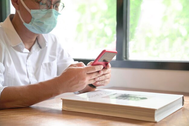 Man with face mask sitting next to windows texting on his phone