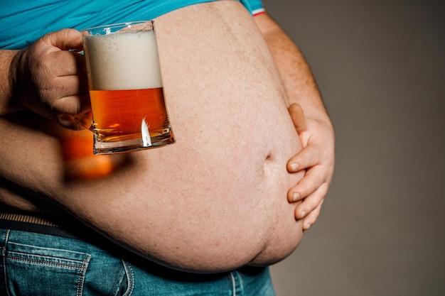 Мужчина с толстым животом держит стакан пива на темном фоне