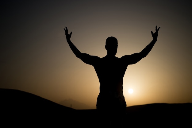 Man with fit silhouette on sunset sky. Sportsman with raised hand on natural landscape. Athlete show power. Workout in summer dusk. Sport, wellness and bodycare concept.