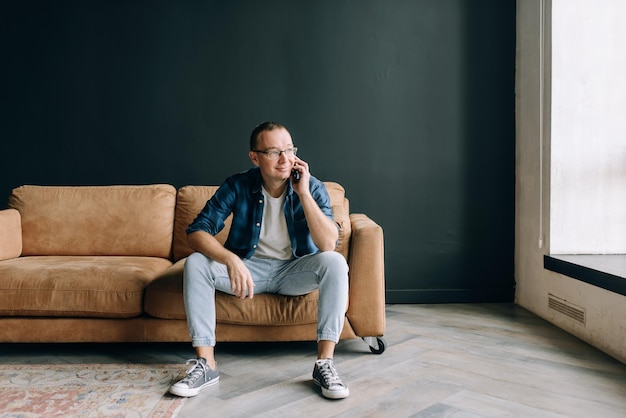 Foto un uomo con gli occhiali in abiti casual seduto sul divano sorridente parlando al telefono