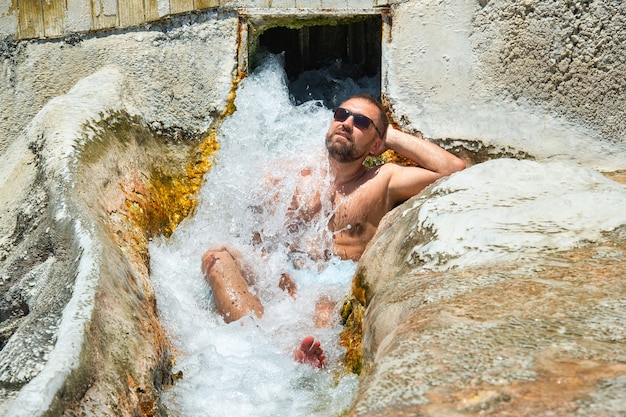 A man with glasses sits under a waterfall of healing water with\
thermal springs in pamukkale.turkey