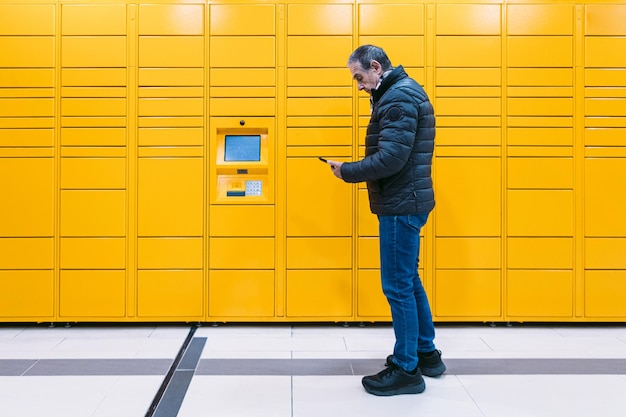 Man with gray coat looks at his cell phone to pick up a package from the locker. messaging concept, compare online, e-commerce and packages