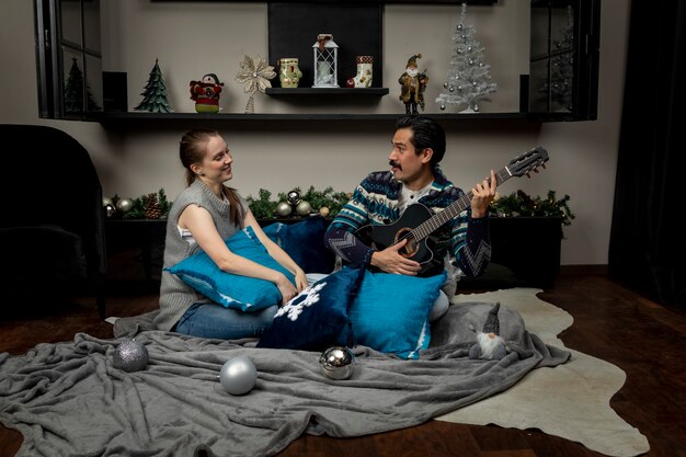 Man with guitar singing to his girlfriend sitting in his living room with pillows on a christmas night