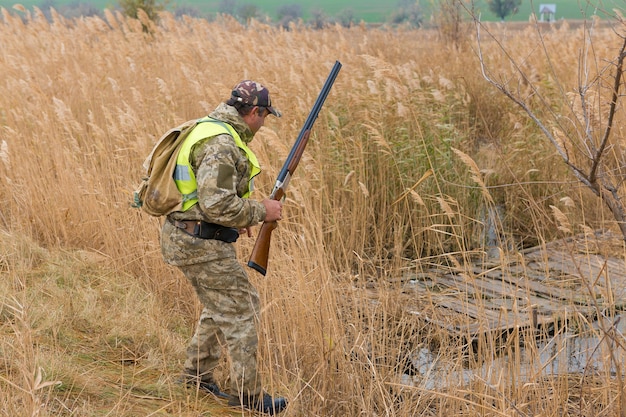 A man with a gun in his hands and an green vest on a pheasant hunt in a wooded area