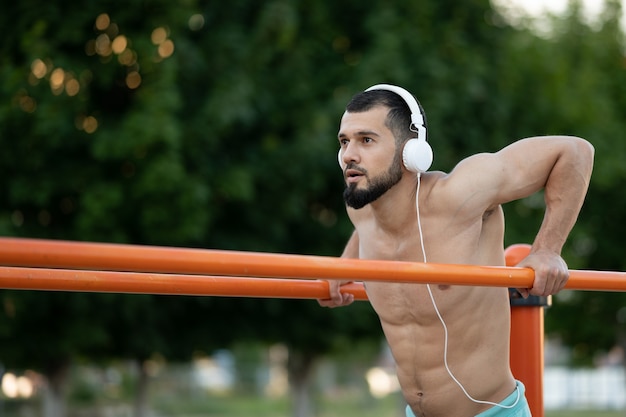 A man with headphones doing push UPS on the bars on the street at the day