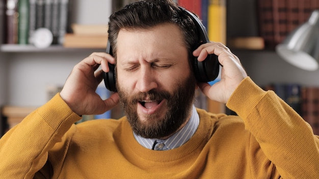 L'uomo con le cuffie ascolta musica e canta. uomo barbuto positivo in cuffie wireless nere tiene la testa con le mani e canta una canzone mentre si gode la musica. colpo medio