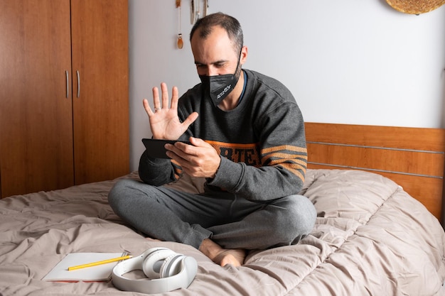 ヘッドフォン、携帯電話、フェイスマスクを持ち、ベッドに座って自宅の部屋に閉じ込められた男