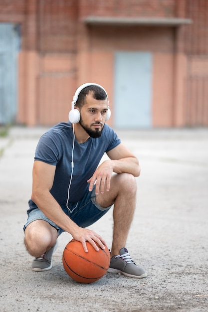 A man with headphones on the street is preparing to play\
basketball, sitting holding a basketball in his hand