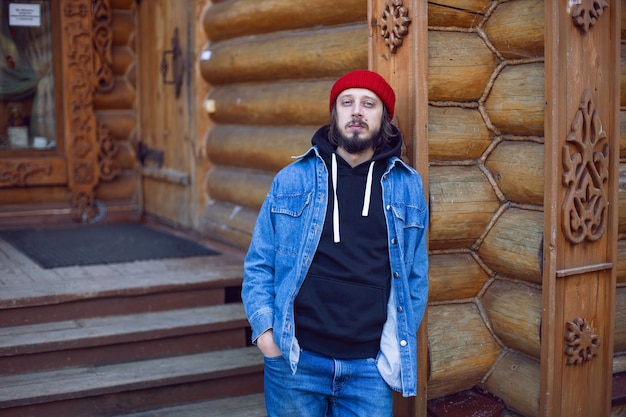 Man with a hipster beard in a red knitted hat and a denim jacket, is stand at wooden house in the woods