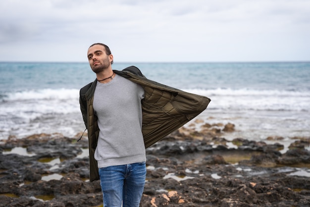 Man with his hands in his pockets stretching his jacket. He is at sea on a cloudy day. He is wearing a green jacket, grey sweater and jeans. Caucasian man with ponytail swinging jacket.