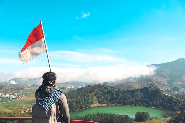A man with Indonesian flag standing on cliff and enjoying the view of nature of lake and mountain in Dieng Indonesia
