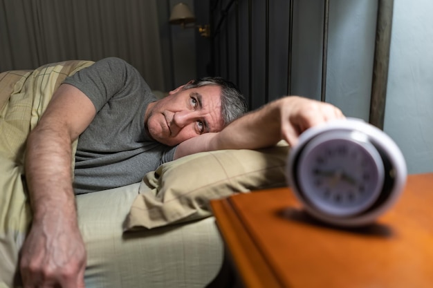 Man with insomnia lying in bed with clock on the bedside table