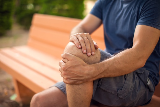 Foto uomo con dolore al ginocchio seduto sulla panchina nel parco. concetto di sanità e medicina