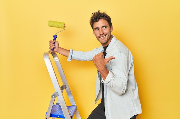 Photo man with ladder and paint roller on yellow points with thumb finger away laughing and carefree