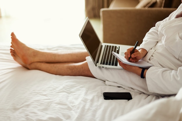 A man with a laptop on the bed the man in the bedroom is lying on a bed with a computer on his lap and a notebook in his hands