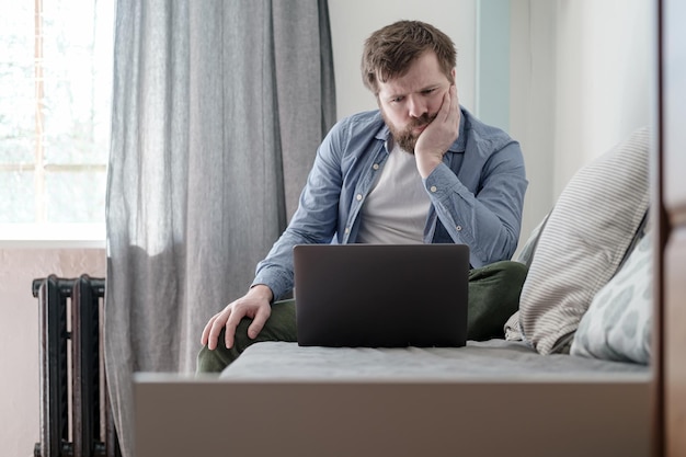 Man with a laptop is bored sitting on a sofa at home