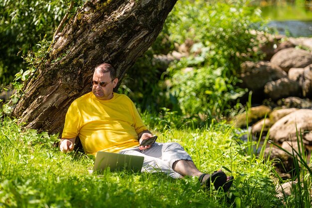 Человек с ноутбуком и смартфоном, работающий снаружи на лугу у реки, концепция офиса под открытым небом