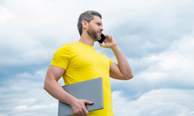 空を背景にスマートフォンで話しているラップトップを持つ男