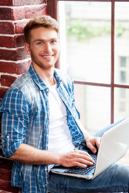 Photo man with laptop. top view of handsome young man working on laptop while sitting on the window sill