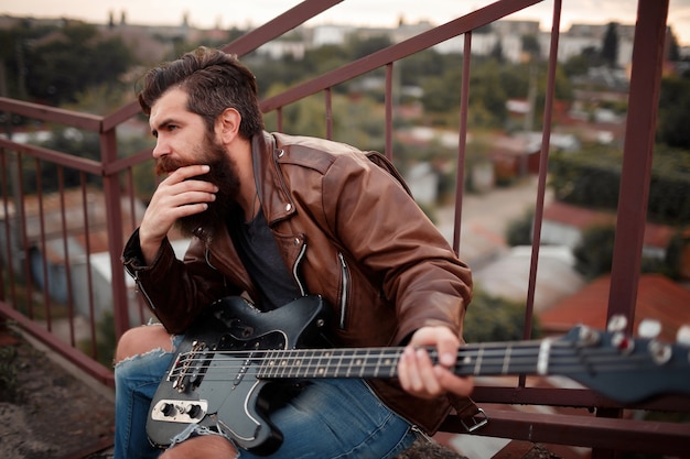 Man with a long beard and gray hair in a brown leather jacket holds an electric guitar in his hand and sits near the stairs against the backdrop of garages