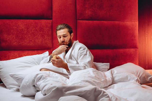 man with a long mustache and beard, wearing a robe, sitting in bed and holding a cream in his hands. Personal care.