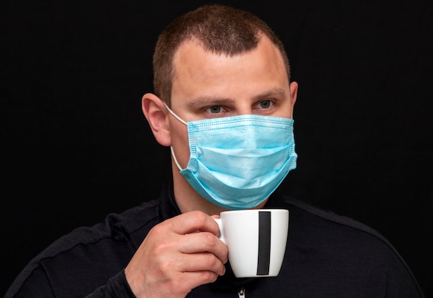 Man with a medical mask drinks coffee on a dark wall