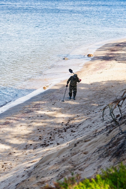 Мужчина с металлоискателем идет у кромки воды песчаного пляжа.