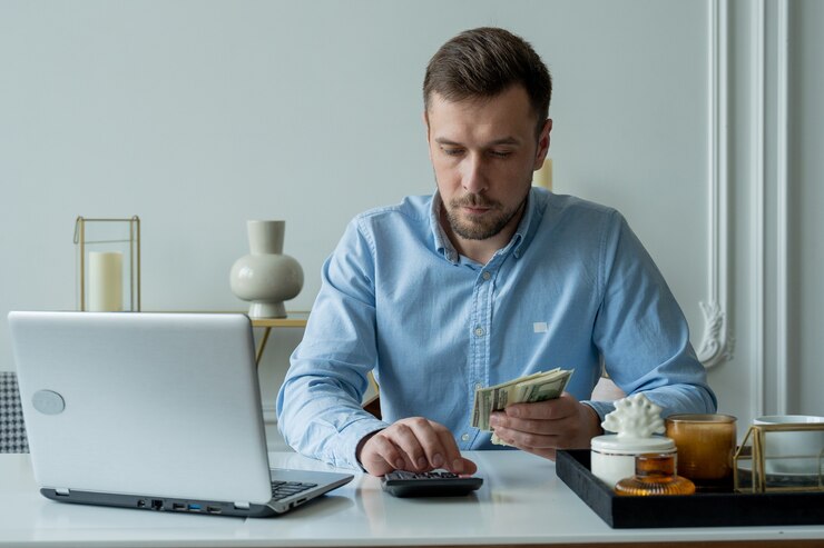 お金と計算機を持った男が請求書をチェックして経費を計算し、自宅のテーブルに座っているクレジット残高を調べます