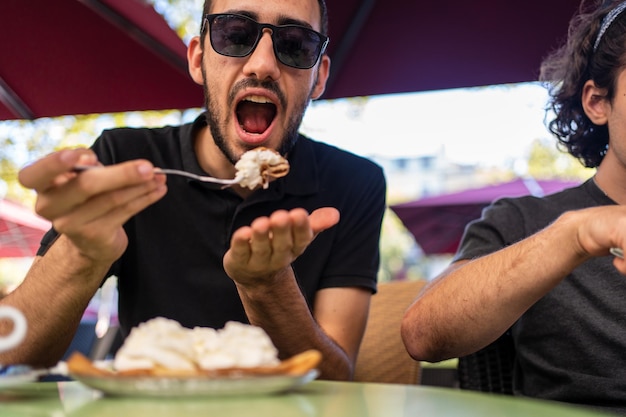 Man with the mouth opened ready to eat a portion of a crepe with cream in a restaurant next to other people