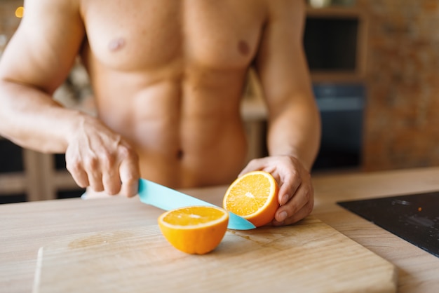 벌거 벗은 몸매를 가진 남자는 부엌에서 오렌지를 자릅니다. 집에서 아침 식사를 준비하는 누드 남성 사람, 옷없이 음식 준비