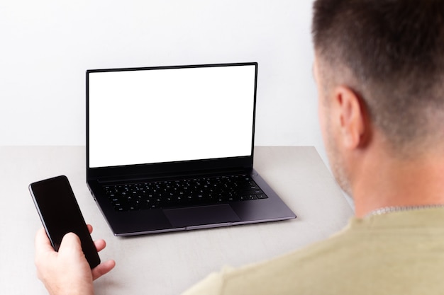 A man with a phone in his hand is sitting in front of a laptop with a white mockup on the monitor, the concept of office work, remote work, marketing, training, coaching