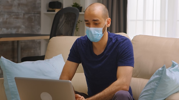 Man with protection mask waving during a video call on laptop. Coronavirus self isolation.