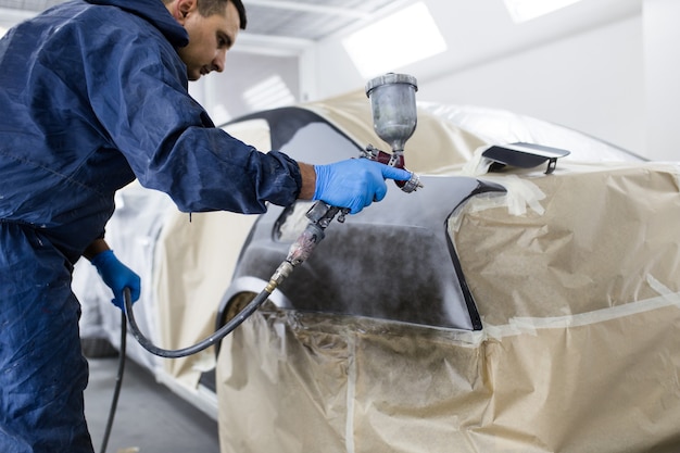 Man with protective clothes and mask painting car using spray compressor. Selective focus.