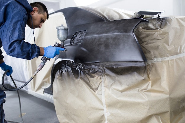 Man with protective clothes and mask painting car using spray compressor. Selective focus.