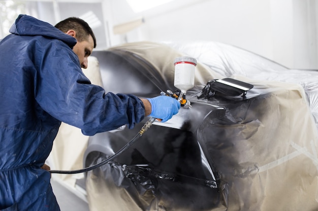 Man with protective clothes and mask painting car using spray compressor. Selective focus.