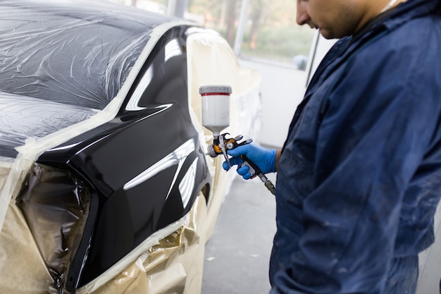 Man with protective clothes and mask painting car using spray compressor. Selective focus.