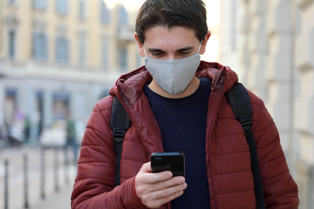 保護マスクを持った男が通りを歩いている間彼のスマートフォンを見る