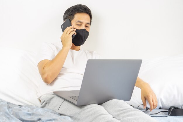 Man with protective mask talking on cell phone with laptop on laps working from home concept.